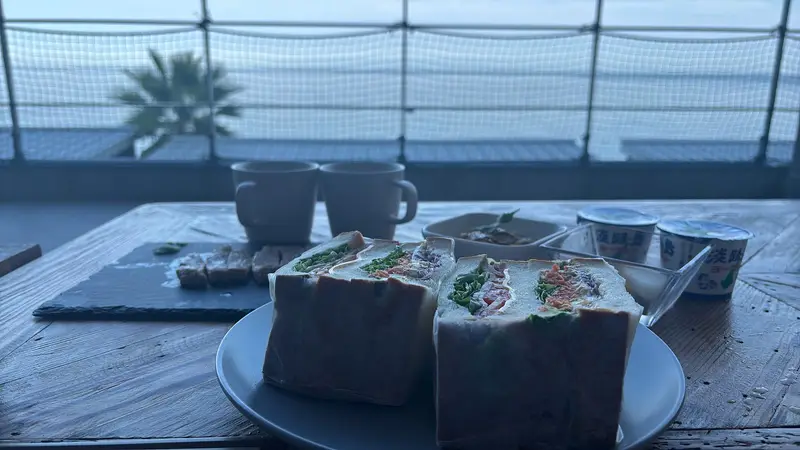 朝のテラスでの朝食。具だくさんのサンドイッチ、淡路島ヨーグルト、湯気の立つコーヒーが二つ、青い朝の海を背に並ぶ