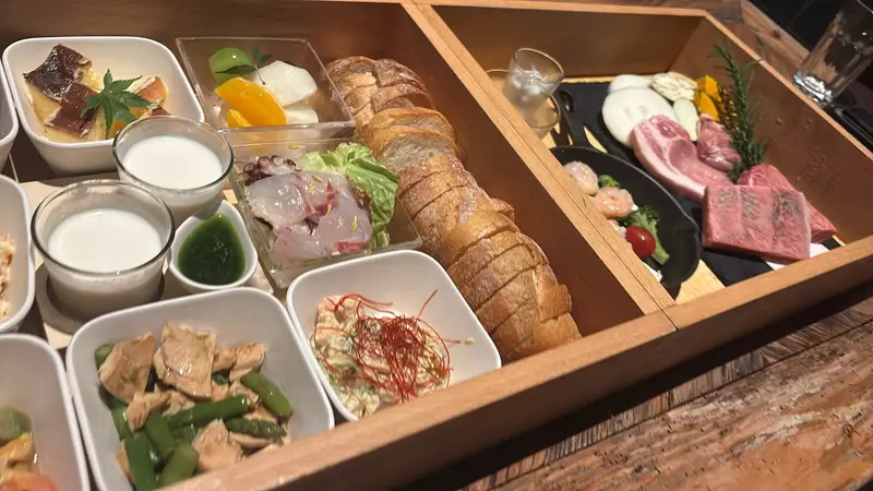 木の三段重に美しく詰められた夕食。前菜の小鉢、白身魚のカルパッチョ、バゲット、焼き用の肉と野菜が並ぶ